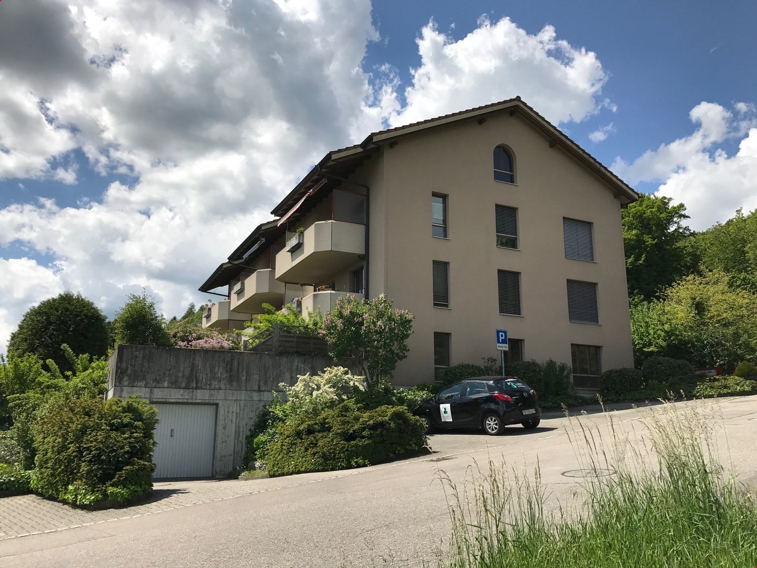 A multi-story apartment building with a parking area in front. The building has a beige exterior with balconies and windows. There are trees and landscaping surrounding the building.
