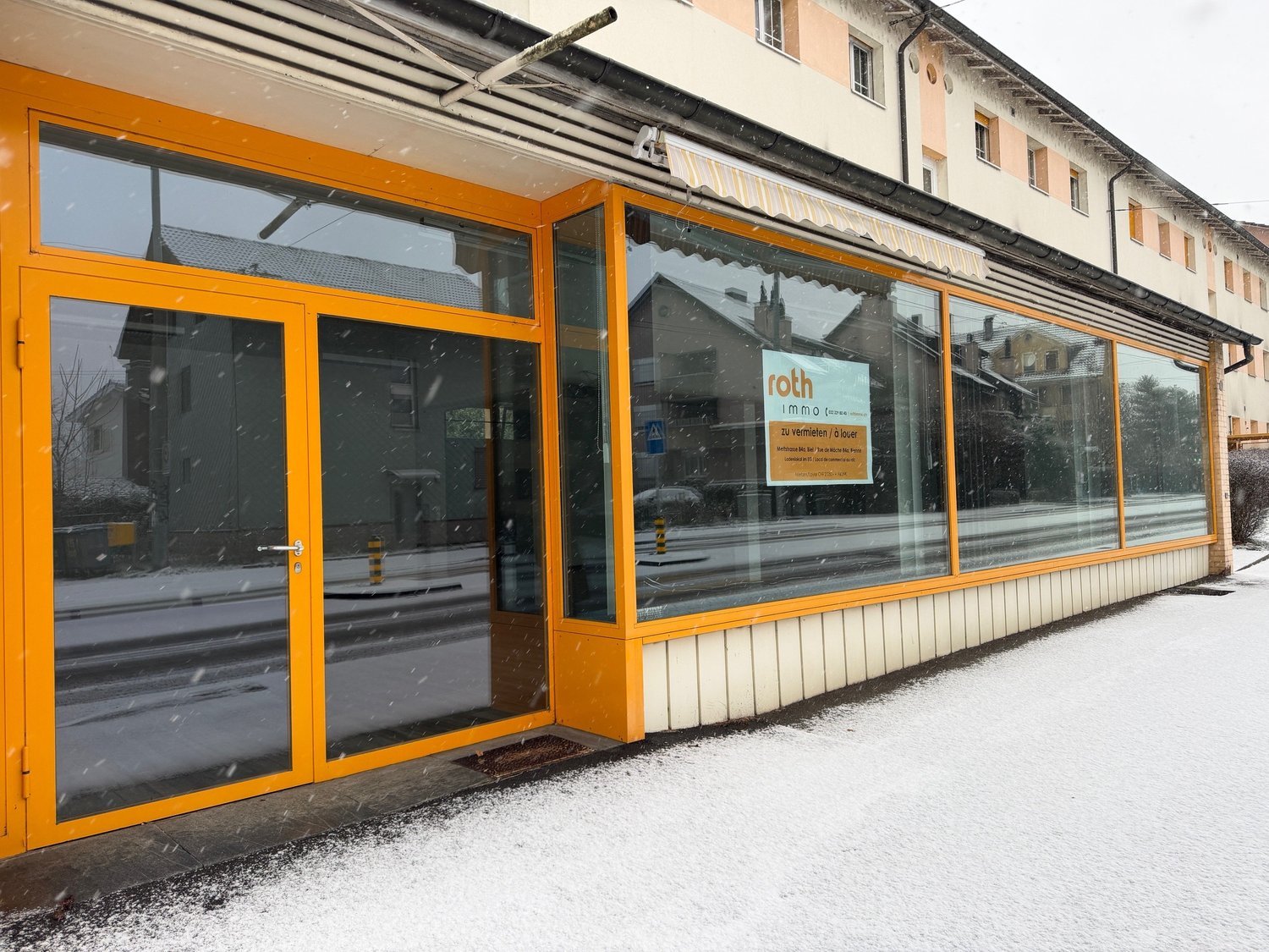 Exterior of a commercial building with glass windows, double-door entrance, signage, and orange frames. Snow on the ground and street.