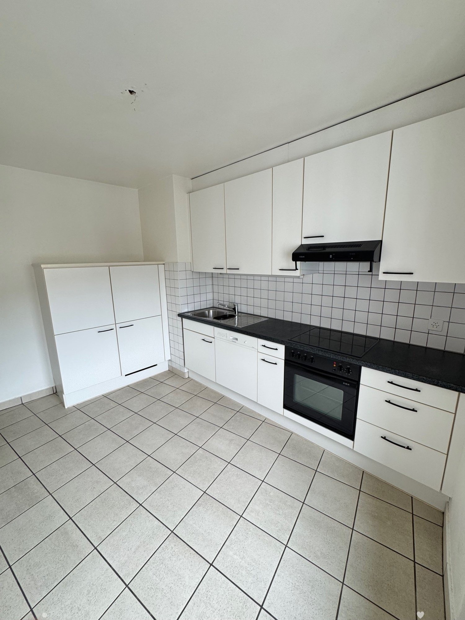 Spacious kitchen with white cabinets, black countertops, tiled backsplash, and built-in appliances including a dishwasher. The floor is covered with gray square tiles.