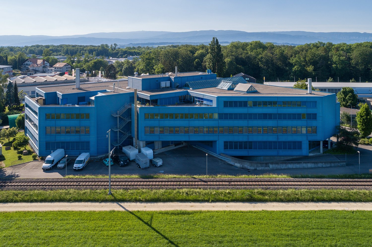 Large blue commercial building with multiple floors, windows, and a rooftop. The building is surrounded by trees and greenery, with a railway track visible in the foreground. The building appears to be located in a scenic, mountainous area.