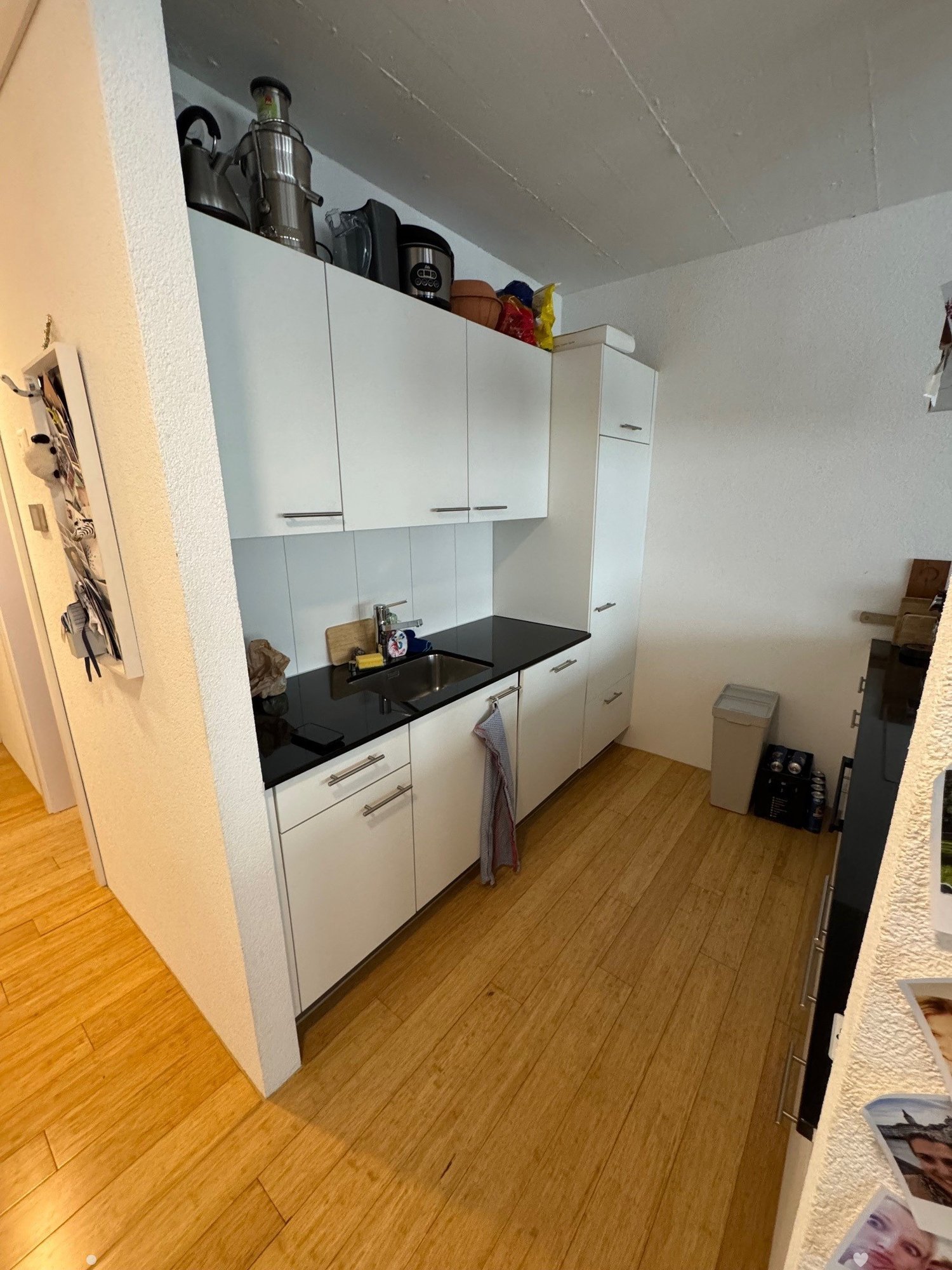 The image shows a small kitchen with white cabinets, a black countertop, and a dishwasher visible on the counter. The floor appears to be made of hardwood or laminate. There are some small appliances and other items stored on top of the cabinets.