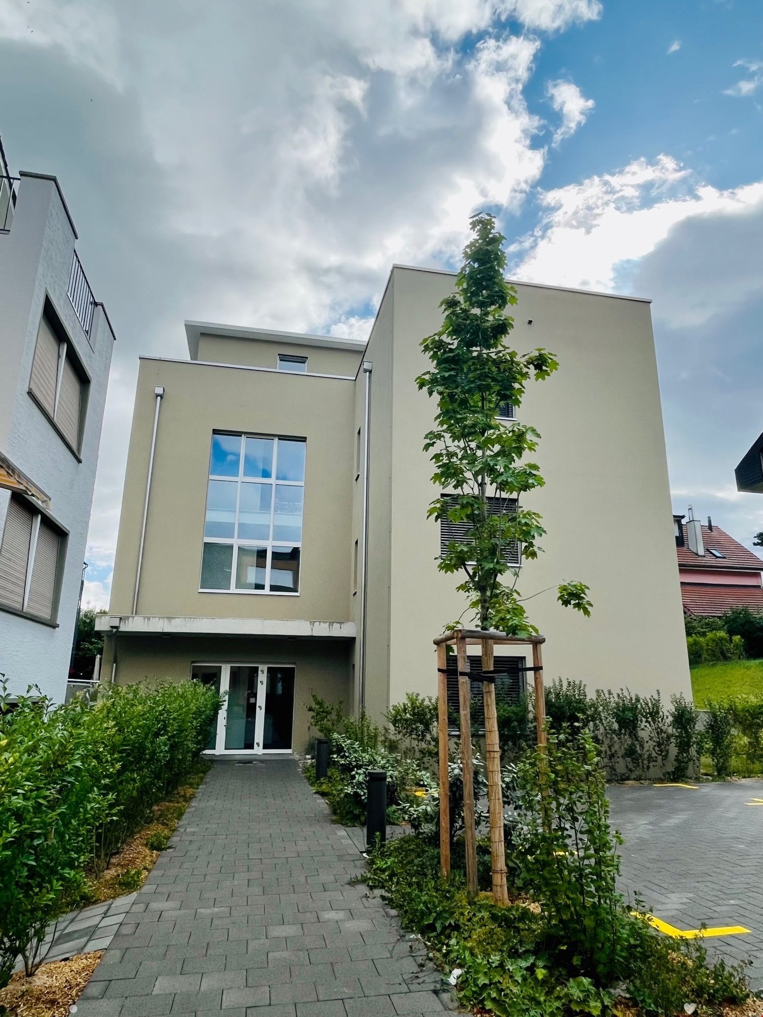 Two-story building with raised ground floor, white walls, large glass windows, front entrance, small plants, and a tree.