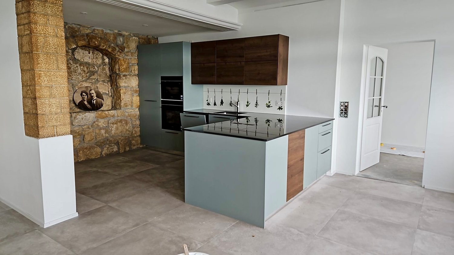 Modern kitchen with island, black countertop, grey cabinetry, stone wall, gas oven, and open door.