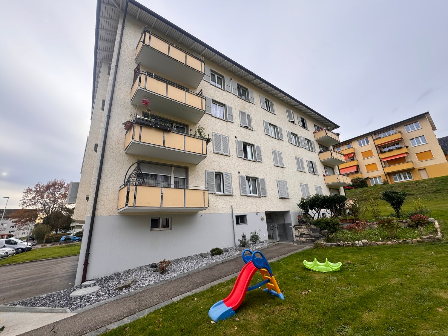 Multi-story building, balconies, small garden area, children's slide, grassy area, several cars parked