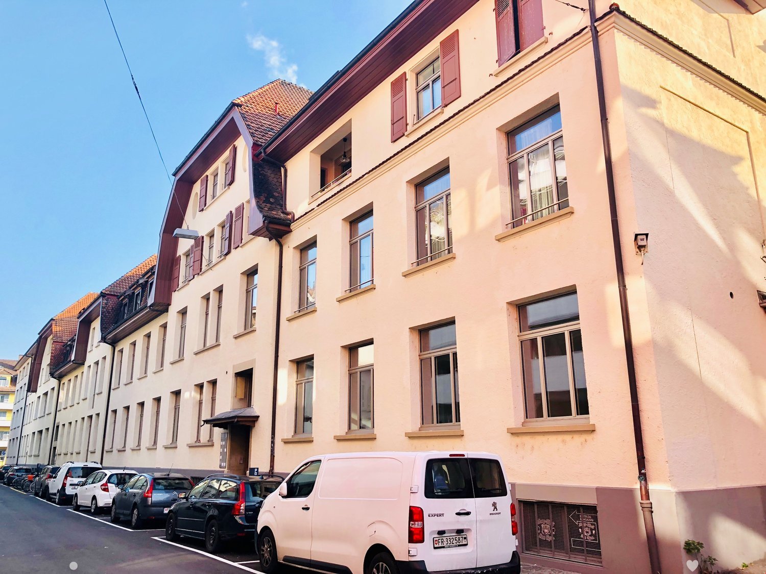 Multi-story building with beige walls, red shutters, many windows, ground floor and upper floors with balconies, white van parked in front.