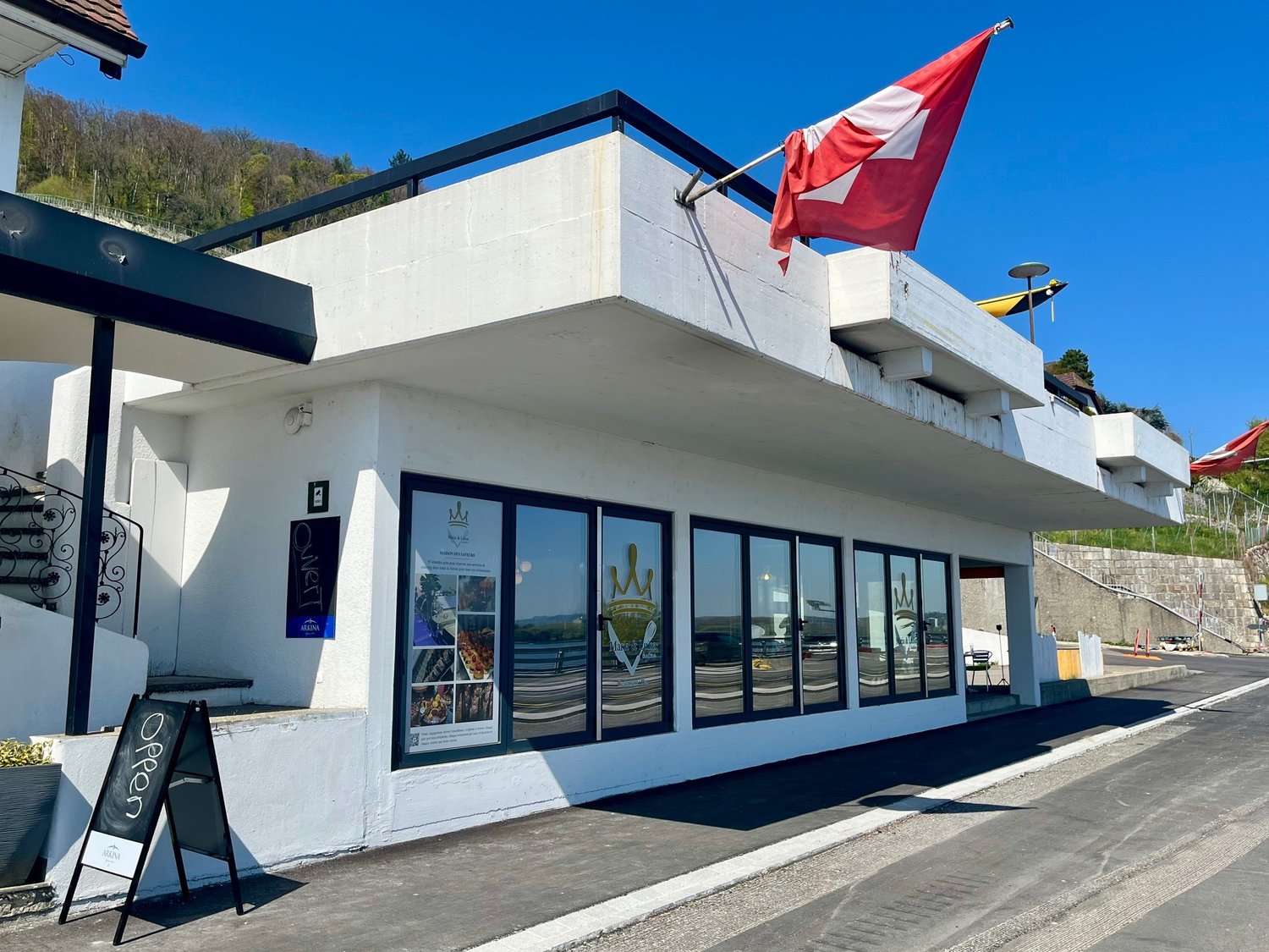 white building, black railing, red and white flag, large glass windows, metal staircase, potted plant, signboard reading 'Open'