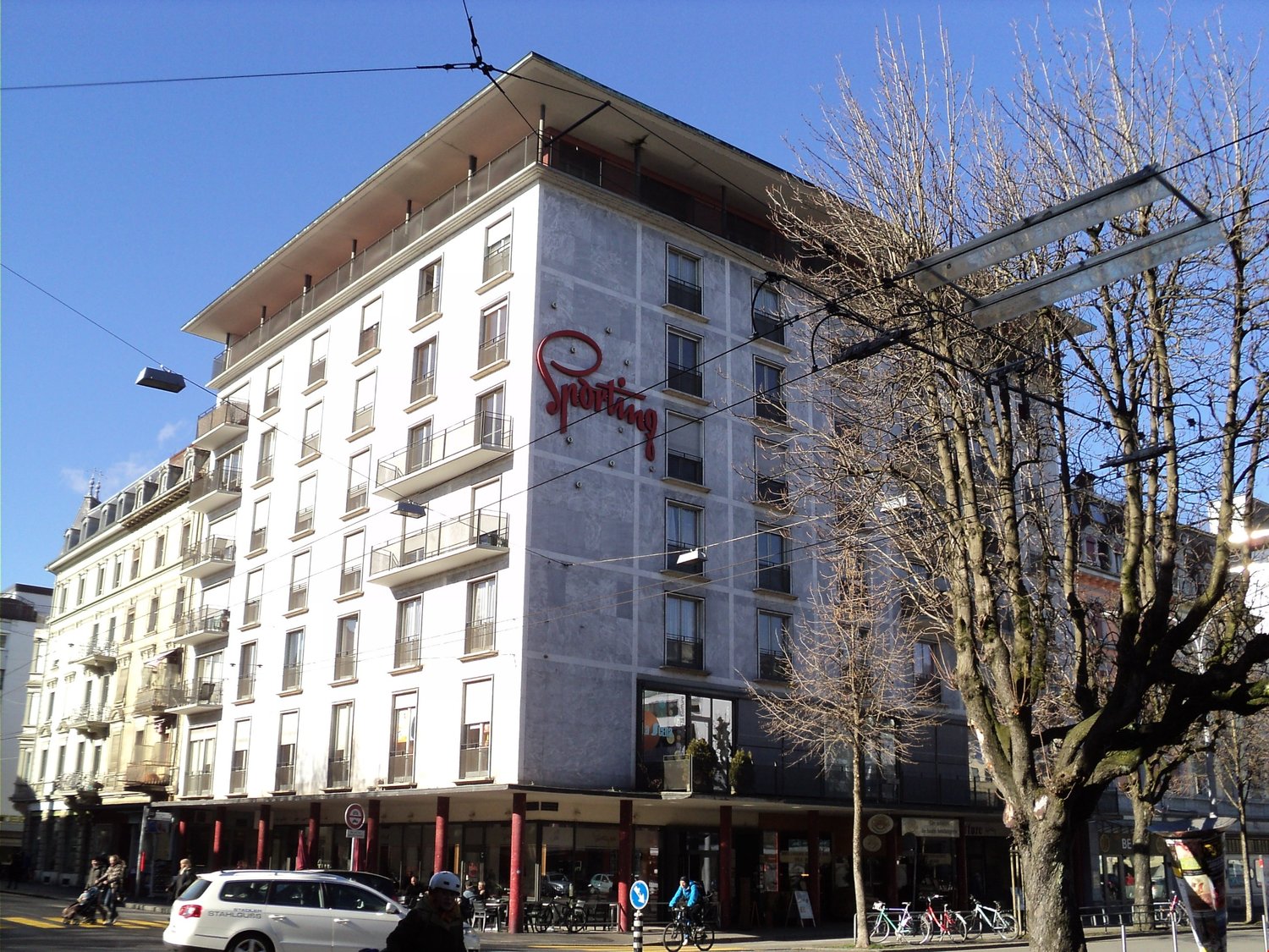 5-story apartment building, red sign 'Sporting', white exterior, glass windows, bicycles parked in front
