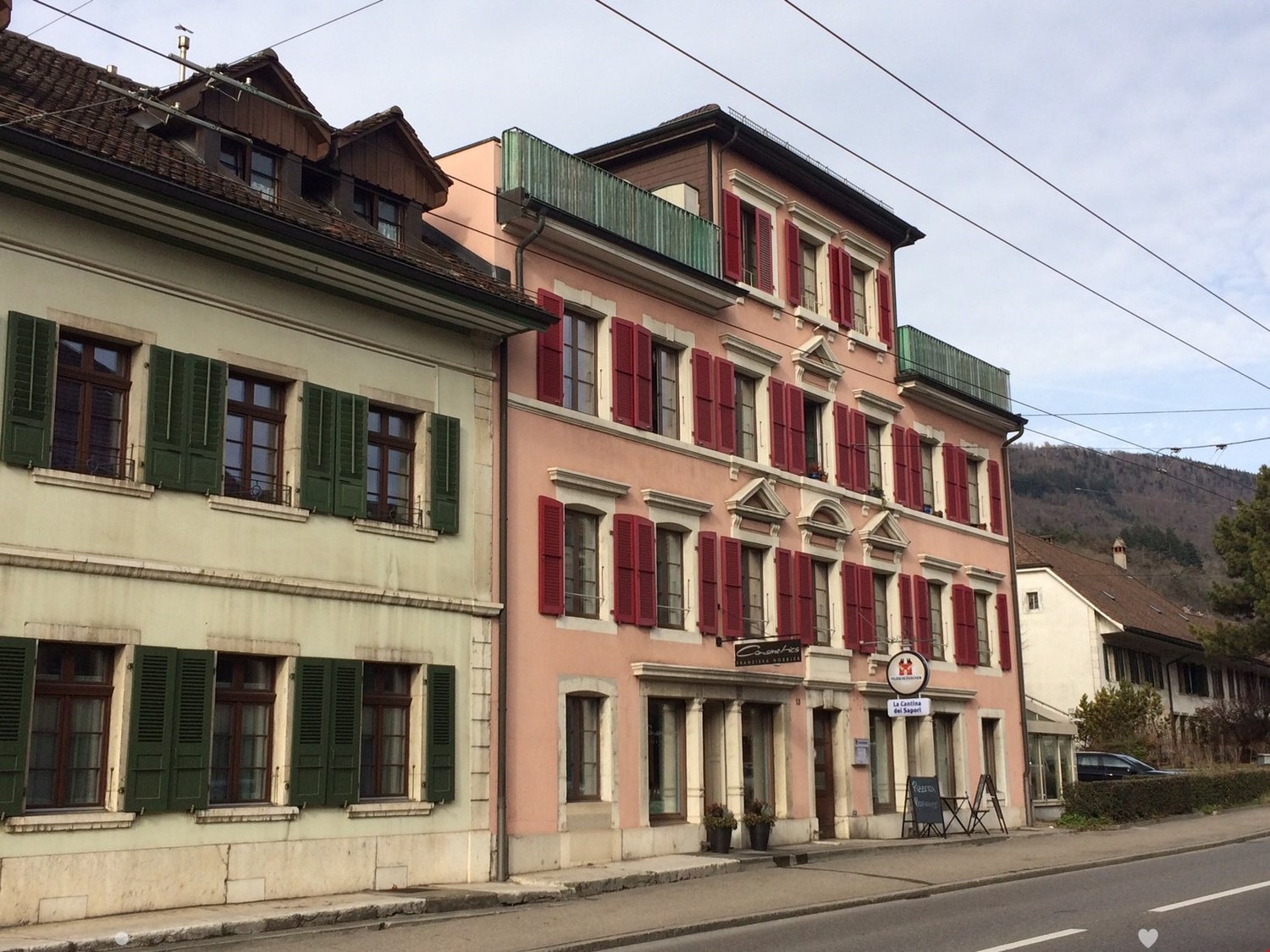 Two-story building, red shutters, green shutters, entrance on ground floor, signage, plants on the sidewalk
