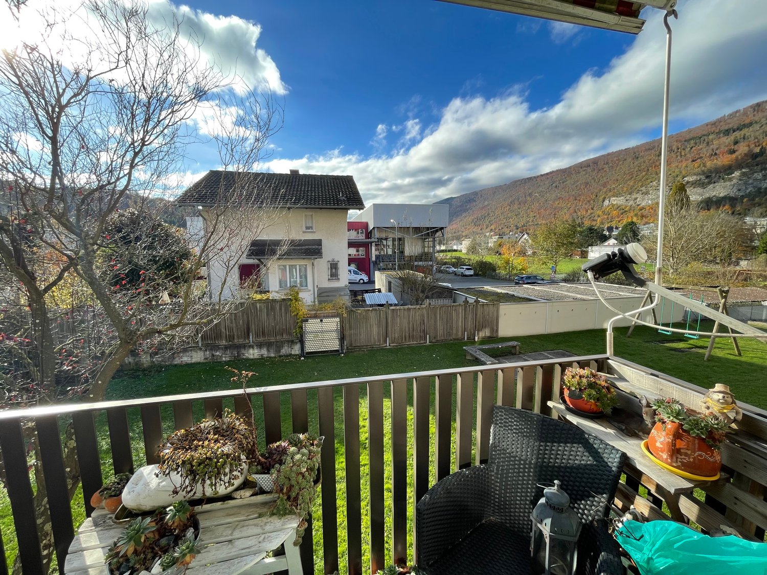 Balcony with fenced railing, small table with plants, wooden bench, swing, lush green grass, a house in the background with trees and parked cars.