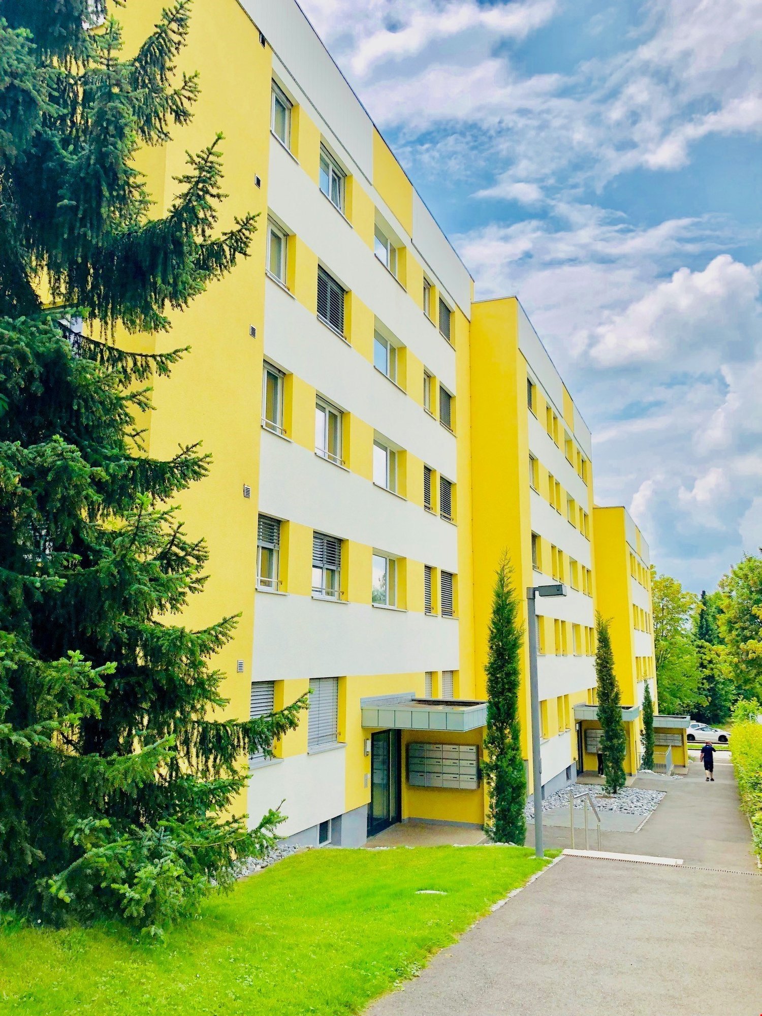 A yellow apartment building with several windows. The building is painted yellow and white. There is a walkway on the right side with a person walking in front of a white car.