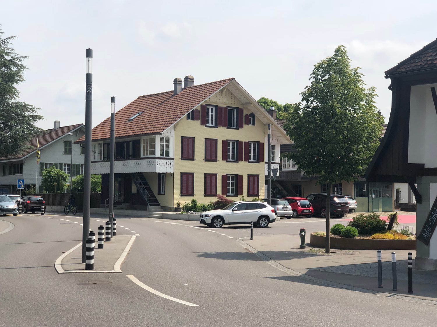 Street scene with a yellow house with red shutters and brown roof, multiple cars parked, sidewalk with street lights, greenery and plants, trees.