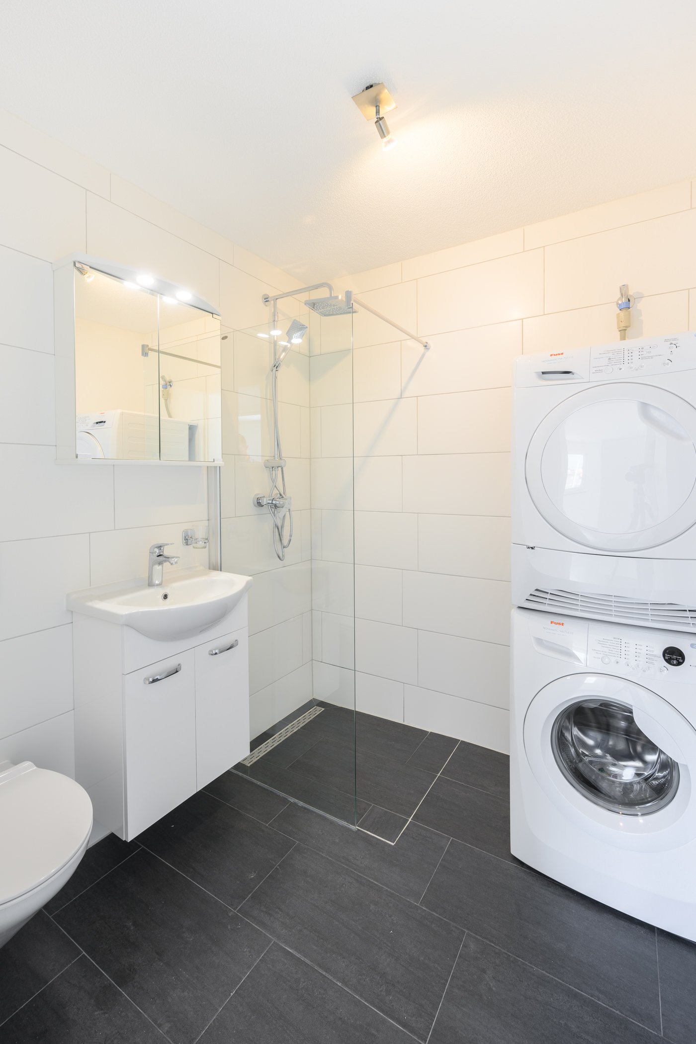 white tiled wall, white tiles on floor, walk-in shower, sink, toilet, mirror above sink, washing machine, dryer