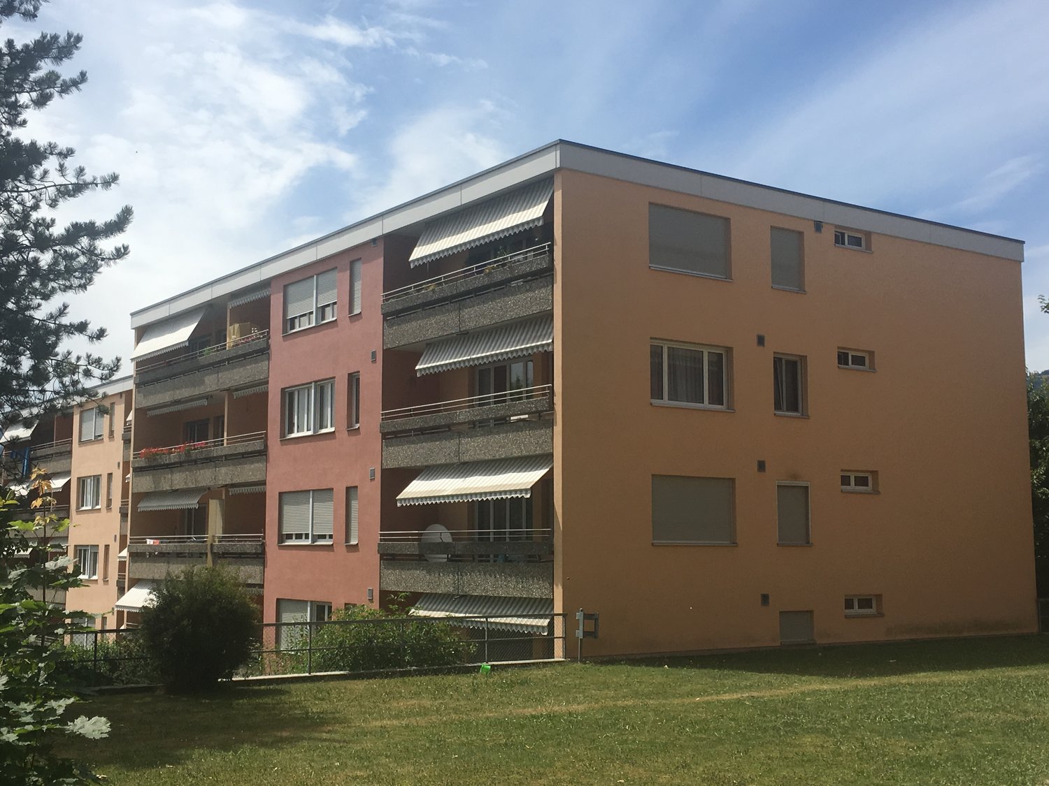 5 story apartment building, beige and red facade, multiple balconies, fenced garden in front