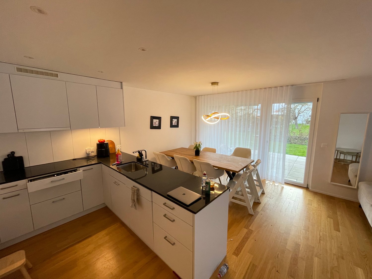 modern kitchen with white cabinets, black countertops, island with sink, dining table, chairs, wooden floor, sliding glass door to patio, pendant light, mirror, framed pictures