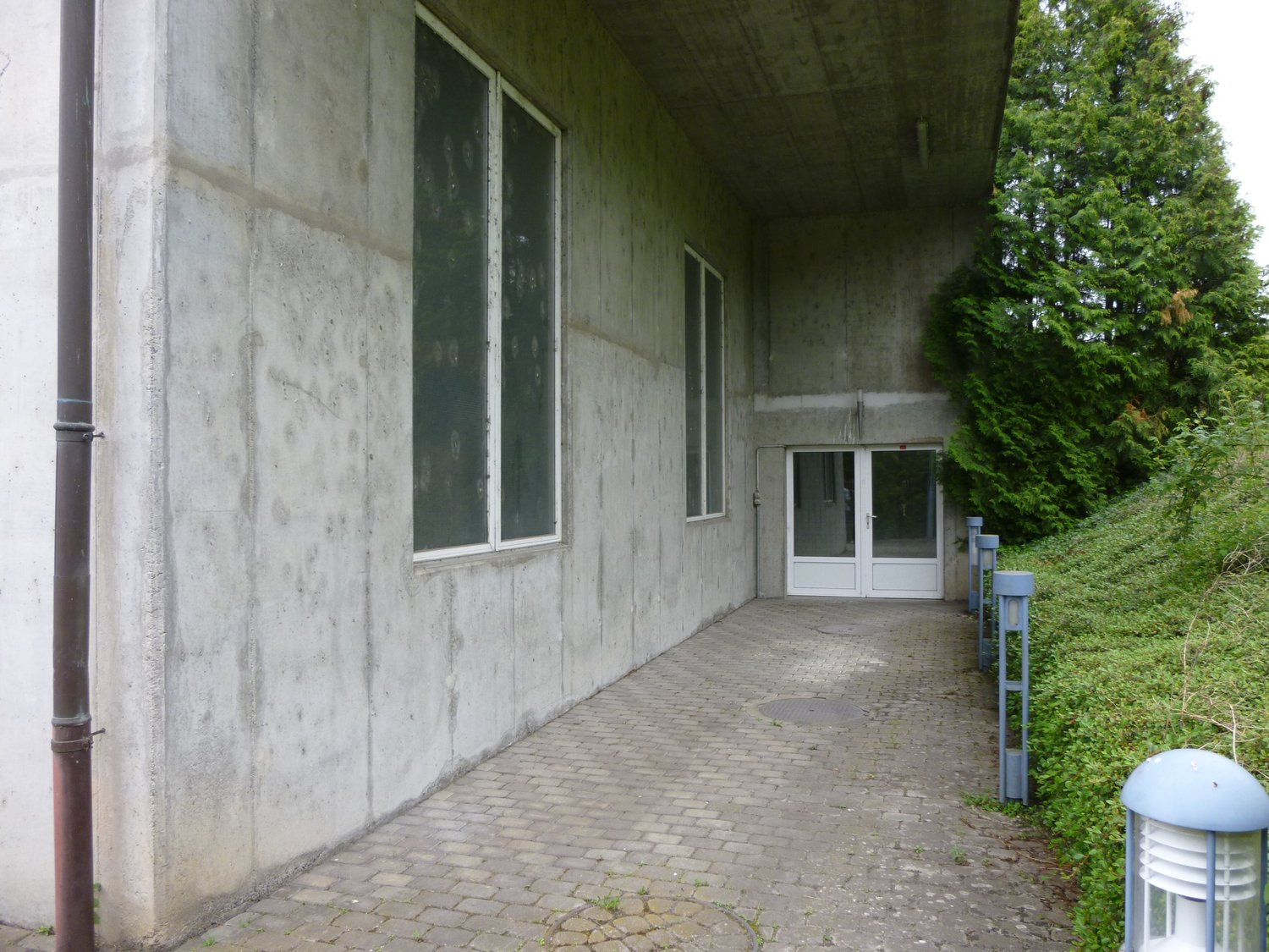 Concrete building with two large windows, brick pathway, white entrance door, and lamp on the right