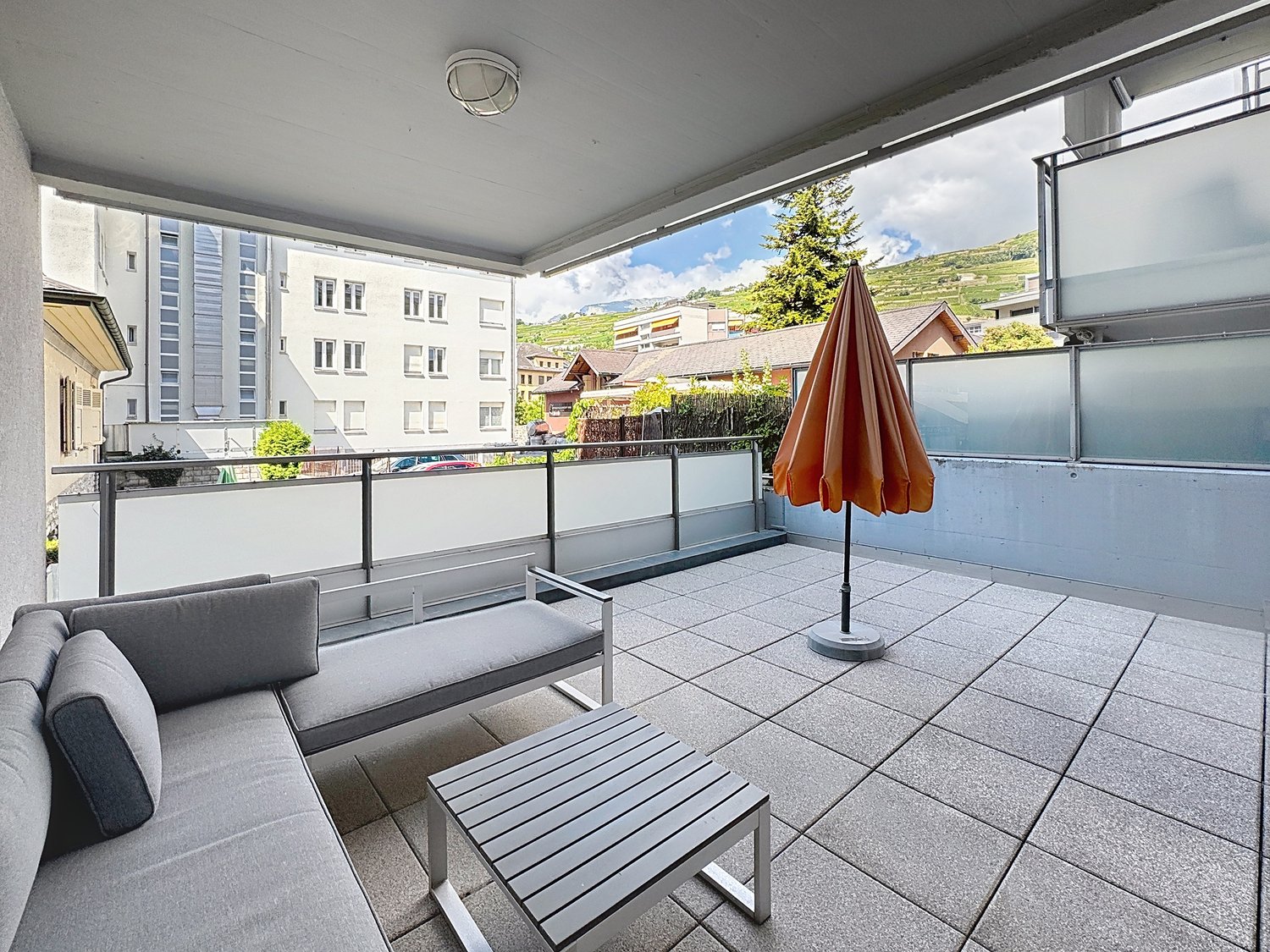 modern balcony with glass railings, metal outdoor furniture, open umbrella, tiled floor, city view