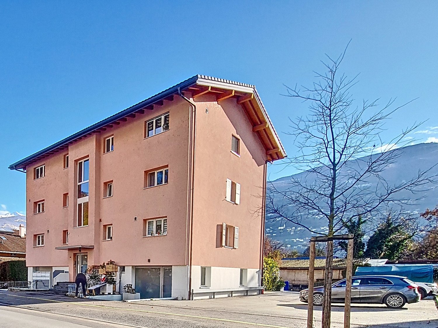 3-story building with 4 apartments, pink facade, brown roof, multiple windows, concrete and brick elements, plants on the first floor, parking area in front, mountains in the background.