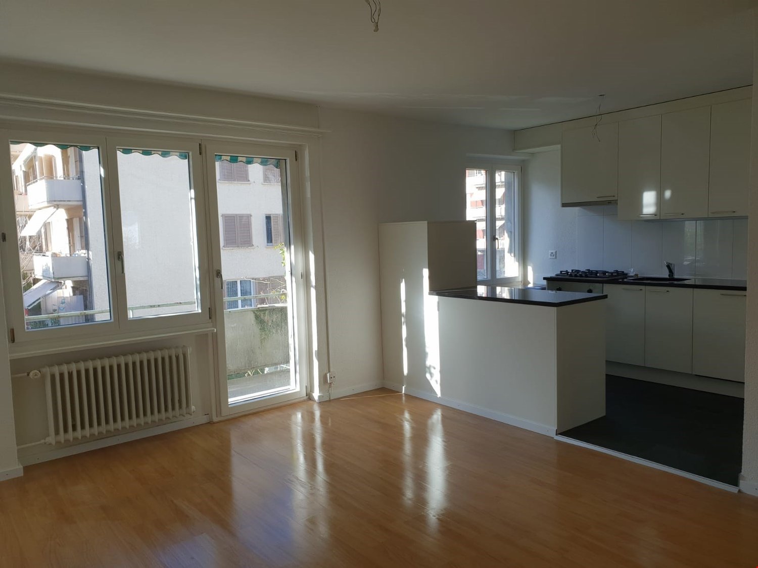 The image shows a spacious open-plan apartment with a living room, kitchen, and balcony. The apartment has large windows that allow natural light to flood the space. The floors are hardwood, and the kitchen features modern white cabinets and appliances. T