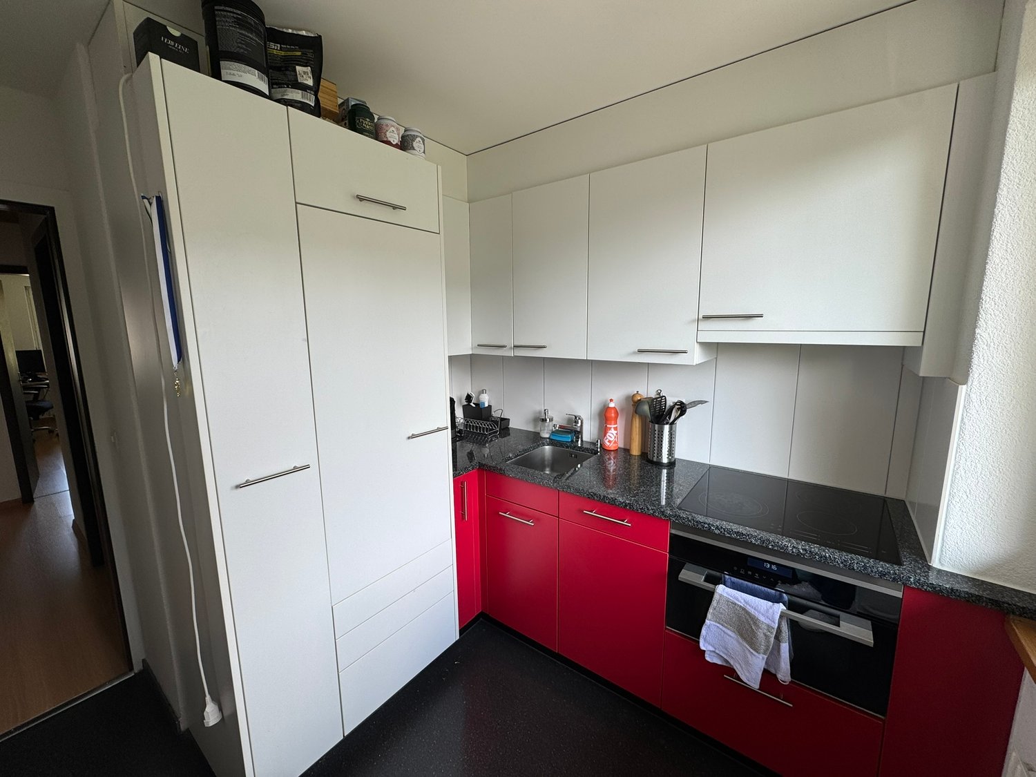 White cabinetry, red base cabinets, black countertops, built-in oven and stove, stainless steel sink and faucet, kitchen tools, and towels.