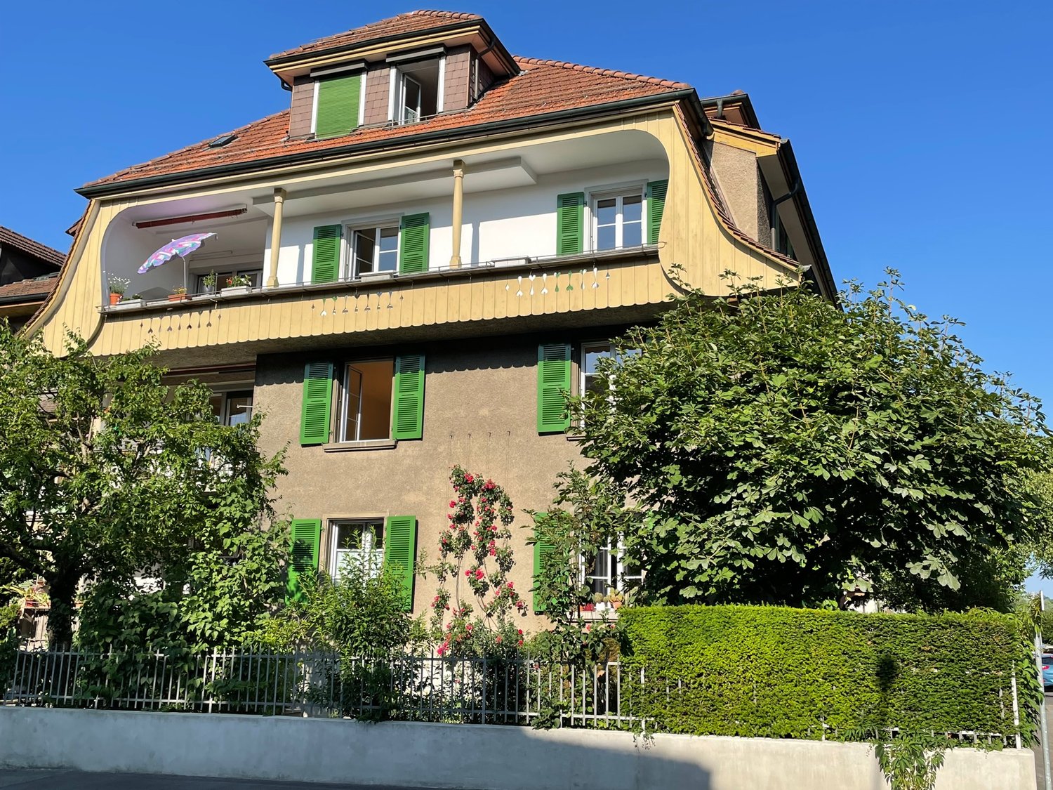 Three story house, beige paint, green shutters, fence, plants, trees