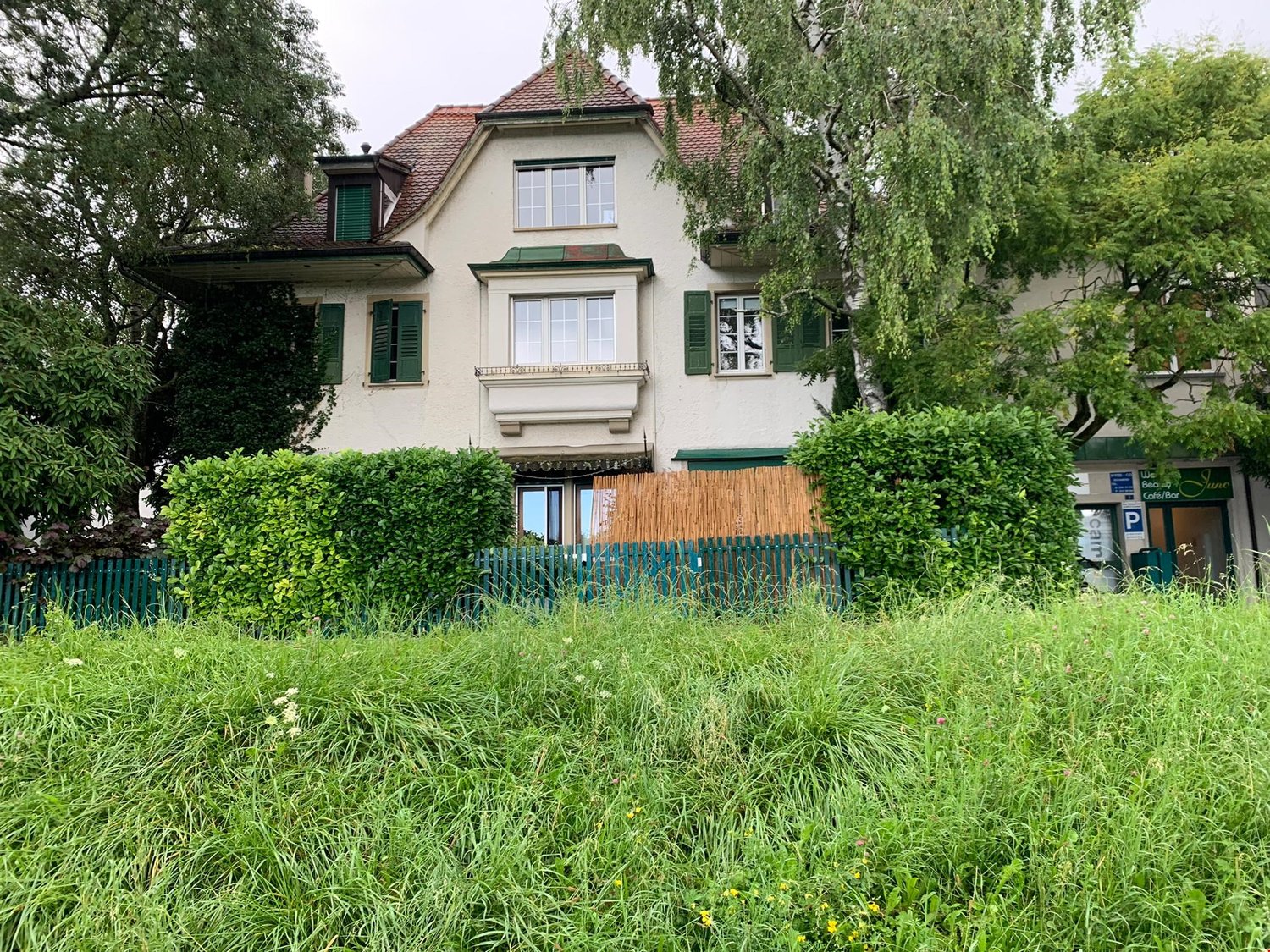 large white villa, red roof, green shutters, covered front yard with shrubs and fence, large windows