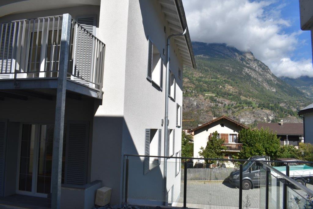white building, glass windows, metal balcony, parked car, surrounding houses, mountain view