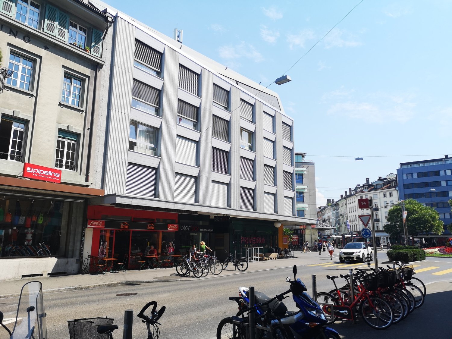 large building with multiple windows, metal railings, ground floor stores, bicycles parked outside, people walking on the sidewalk, parked cars on the street, street lights and trees on the roadside