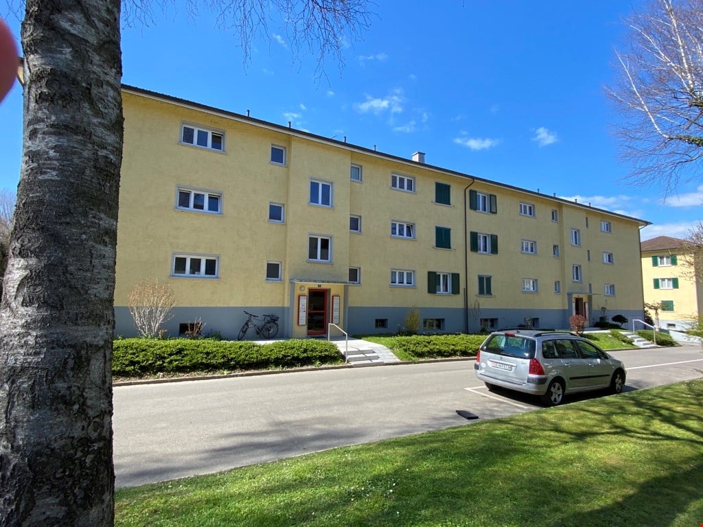 multi-storey residential building, yellow exterior, multiple windows, ground-level entrance with bicycle, parking area with one parked car