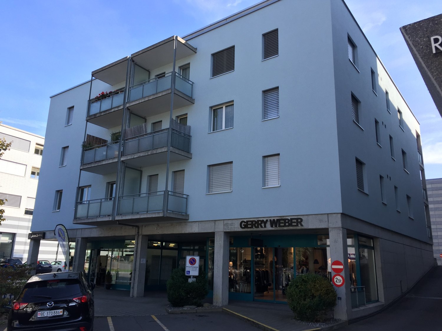 Modern building with 4 floors, white color, balconies, street shop named Gerry Weber, plants on the front