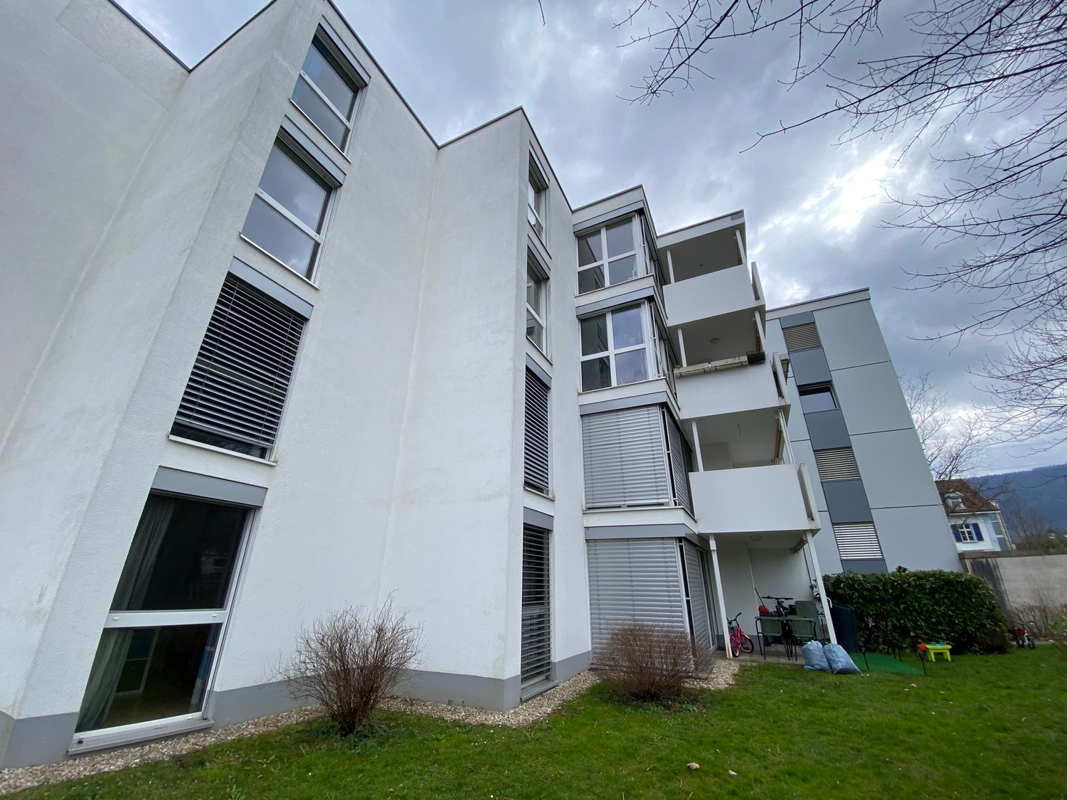 Multi-story apartment building with modern, angular design, large windows, and balconies. The building has a white exterior with gray accents and is surrounded by a grassy area with some landscaping.