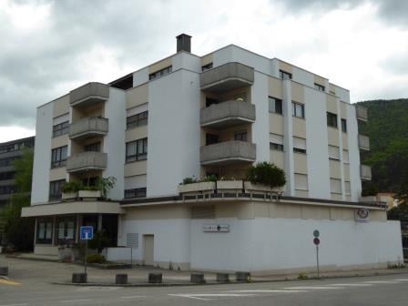 A multi-story apartment building with white exterior walls, balconies, and a few trees or plants visible on the balconies.