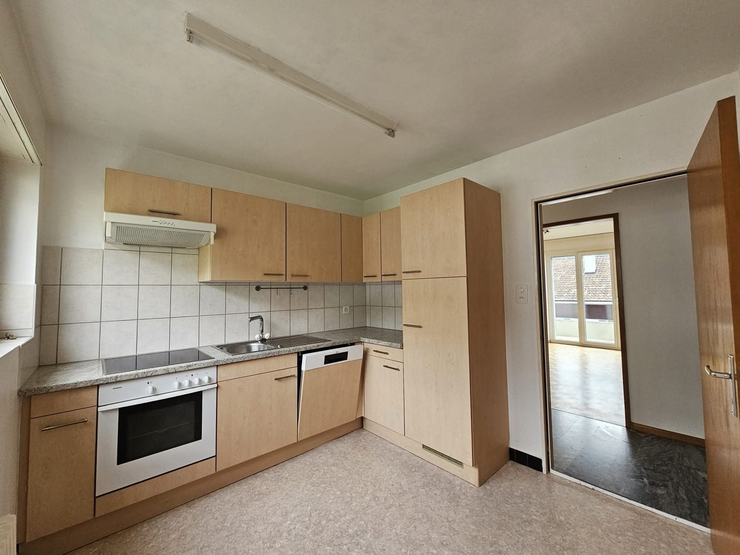 A kitchen with a gas stove, oven, sink, dishwasher, and countertop. Wooden cabinets and countertops are on the walls, and a power outlet is mounted on the wall. There is an entrance to the left side, and a door with a metal handle is open.