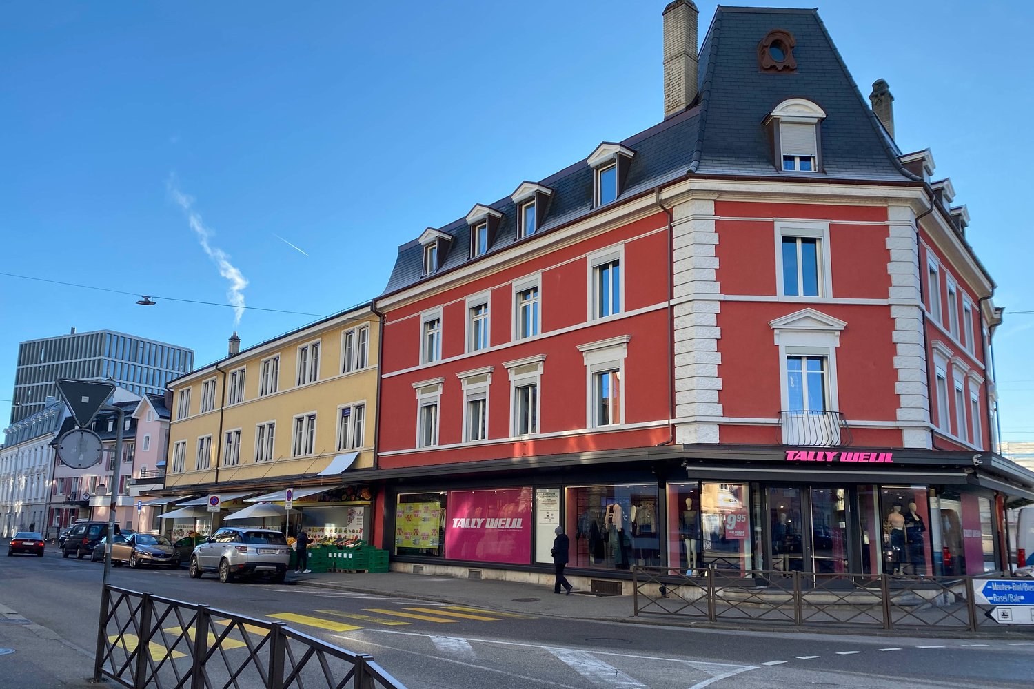 Three-story building with red facade, white trim, multiple windows, storefronts, greenery and cars on the street.