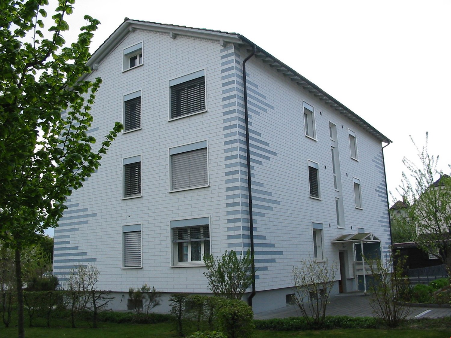 3-story white building, windows with shutters, chimney, entry area with canopy, greenery and plants surrounding the building