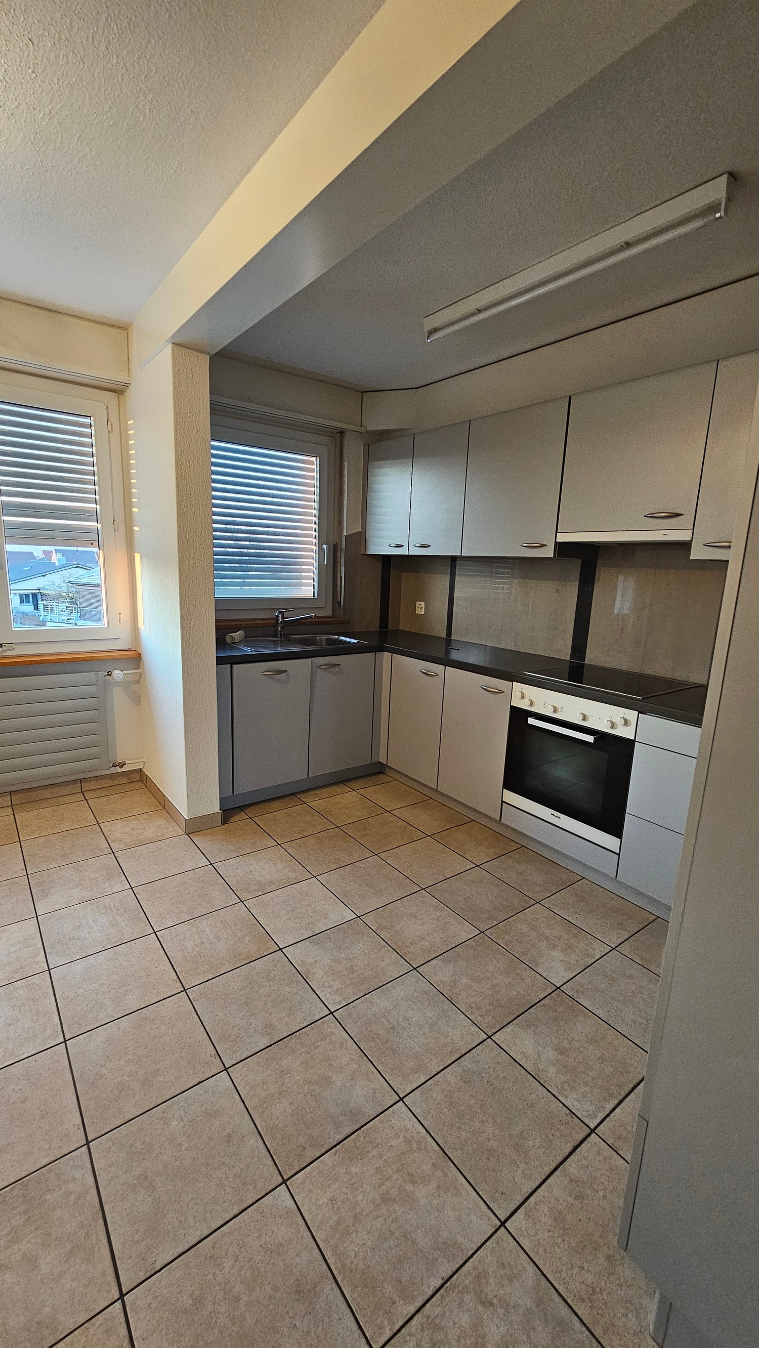 Kitchen with tiled flooring, a sink with faucet, a black countertop, built-in appliances including an oven and a dishwasher, white cabinets, and windows with blinds.