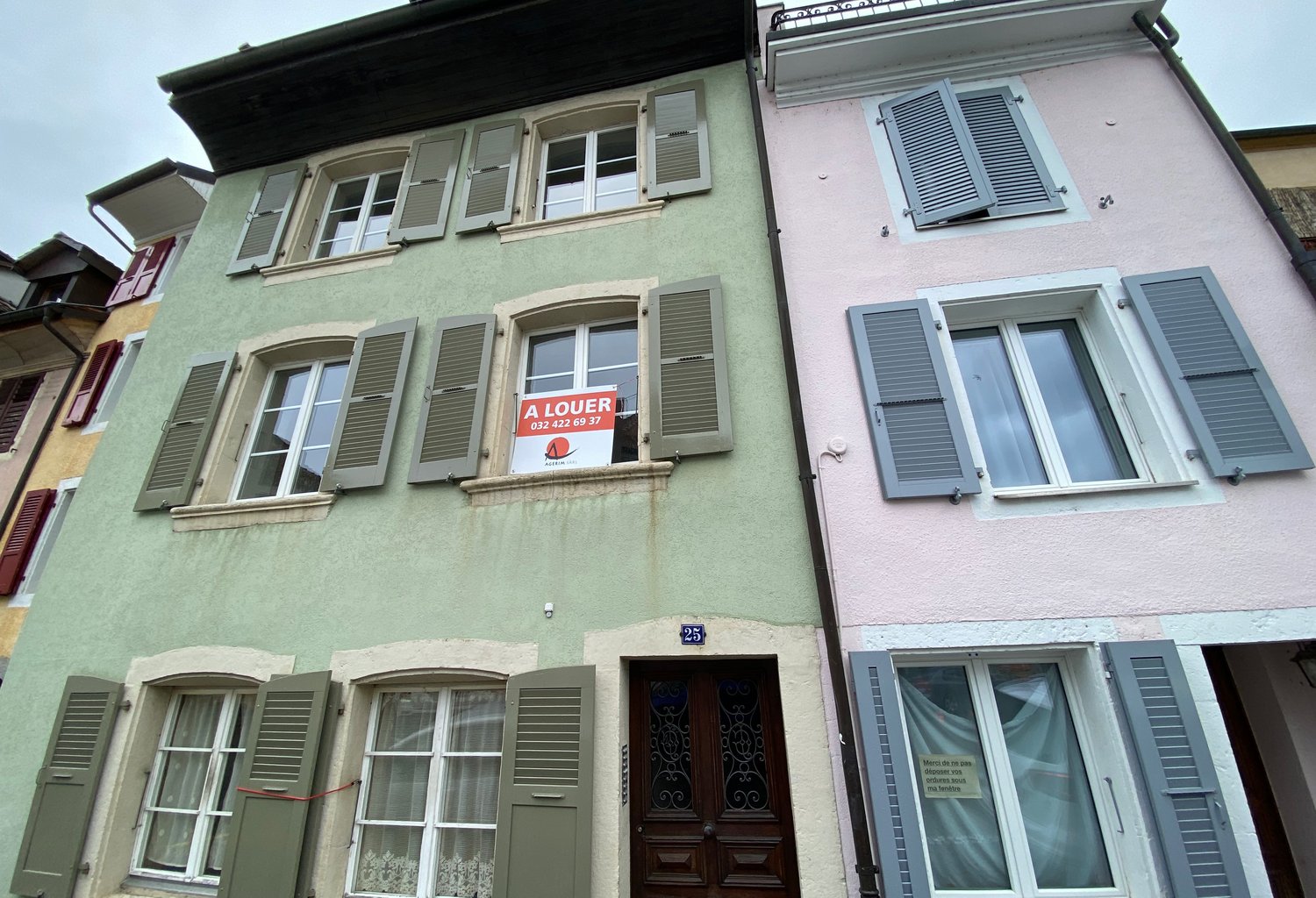 Two-story house, green and pink exterior, multiple windows, wooden door, shutters, sign indicating it is for rent