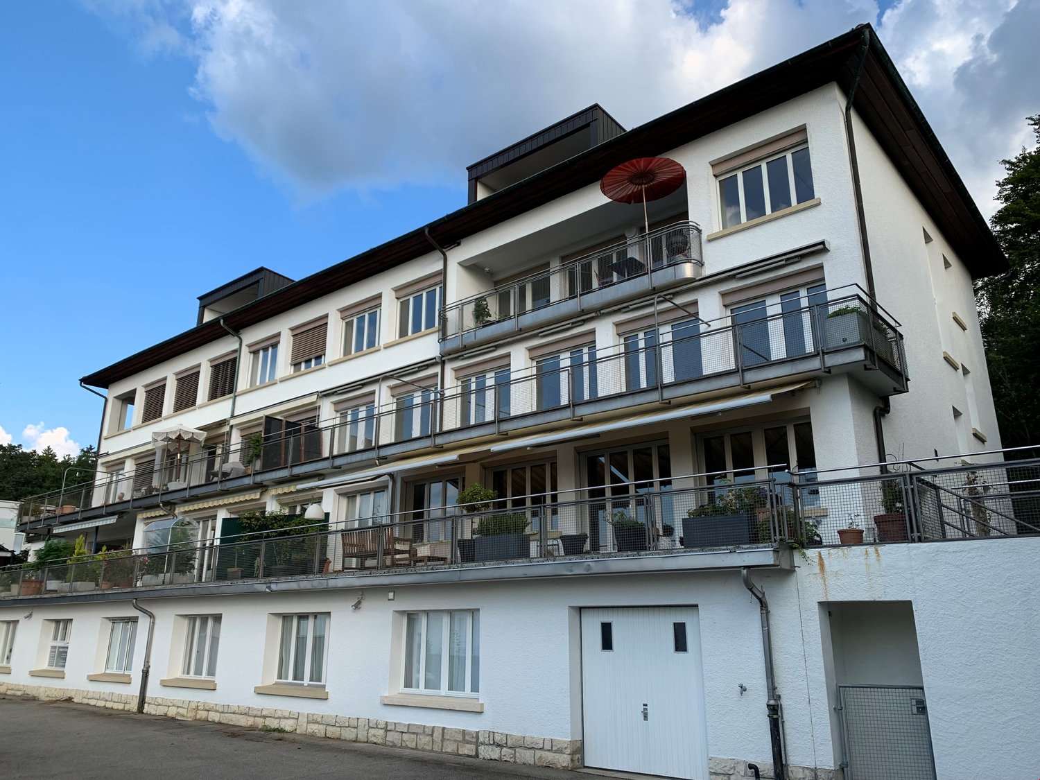 multi-story building, white exterior, multiple balconies with metal railings, green plants in pots, red umbrella, windows with white frames