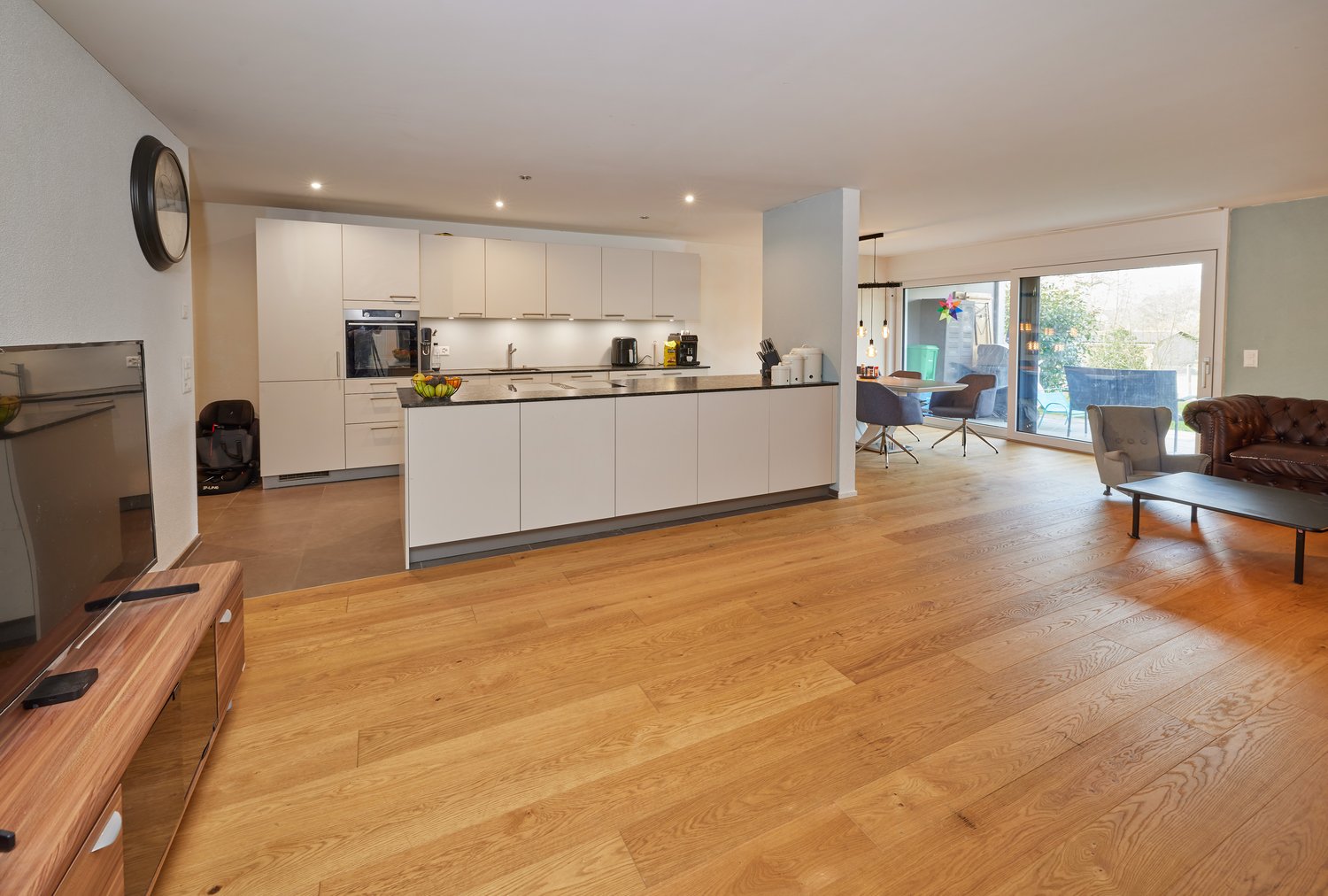 Open plan kitchen and living room, wooden floor, white cabinets, large windows, dining table, couch, TV