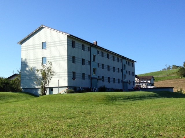 Multi-unit building, white exterior, multiple windows, set on a hill with grassy field