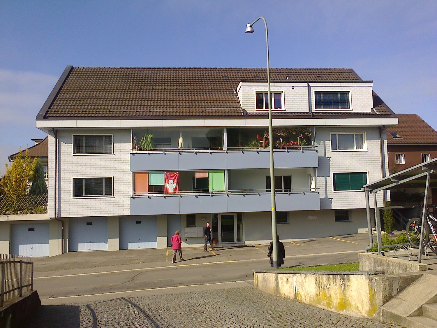 3-story apartment building with white siding, tiled roof, and balconies on the upper floors. There are several people visible on the balconies and walking on the ground floor.