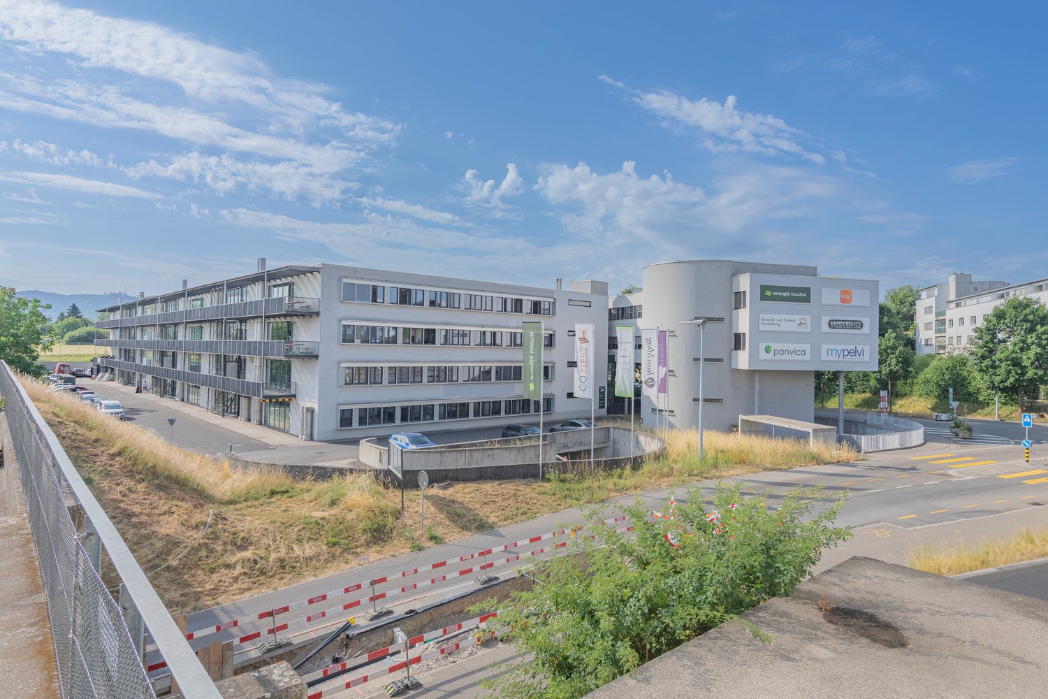 Multi-story office building with glass windows, surrounded by parking spaces and landscaping. The building has signage for various companies like Panvica and Mypelvi.