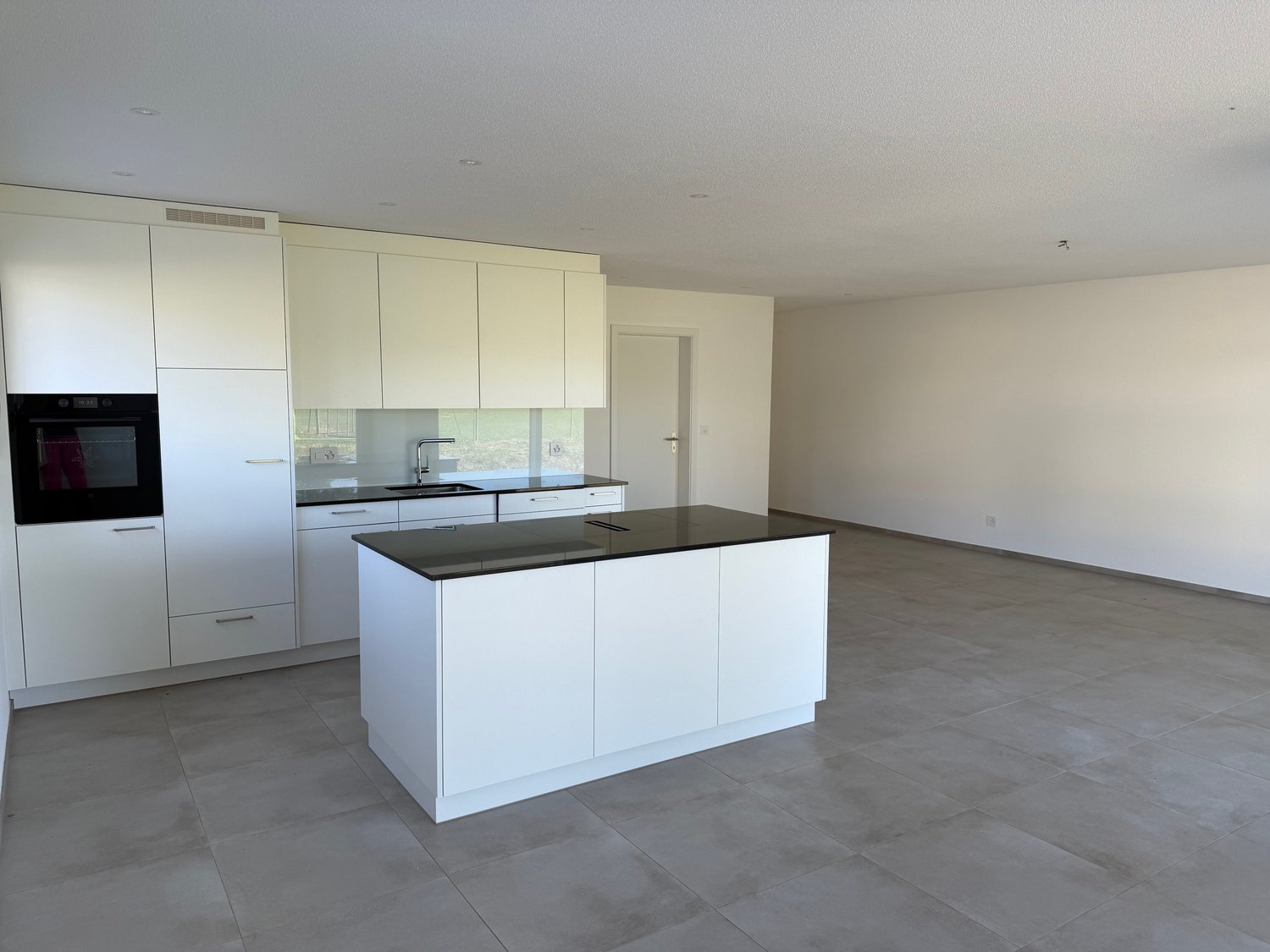 Spacious open-plan kitchen with white cabinets, black countertops, and a central island. The kitchen is equipped with a dishwasher and appears to be part of a larger living space with a tiled floor.