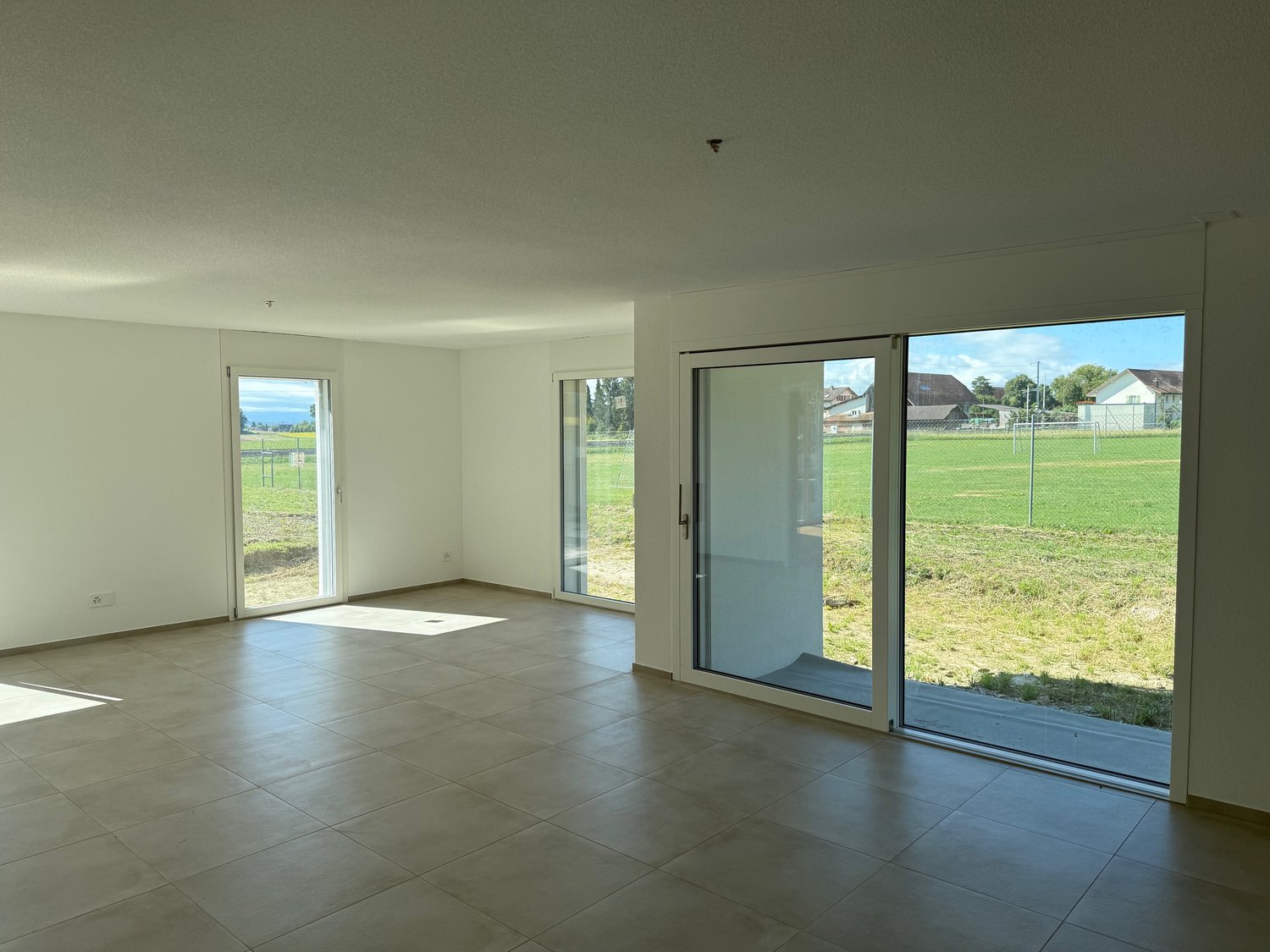 Spacious open-plan living room with large windows and sliding glass doors overlooking a grassy field. The room has a tiled floor and neutral-colored walls, creating a bright and airy atmosphere.