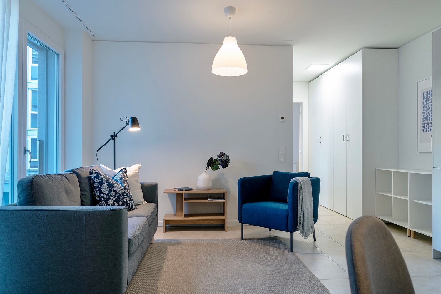 Spacious living room with large windows, gray sofa, blue armchair, wooden side table, and a pendant light fixture. The room has a minimalist and modern design aesthetic.