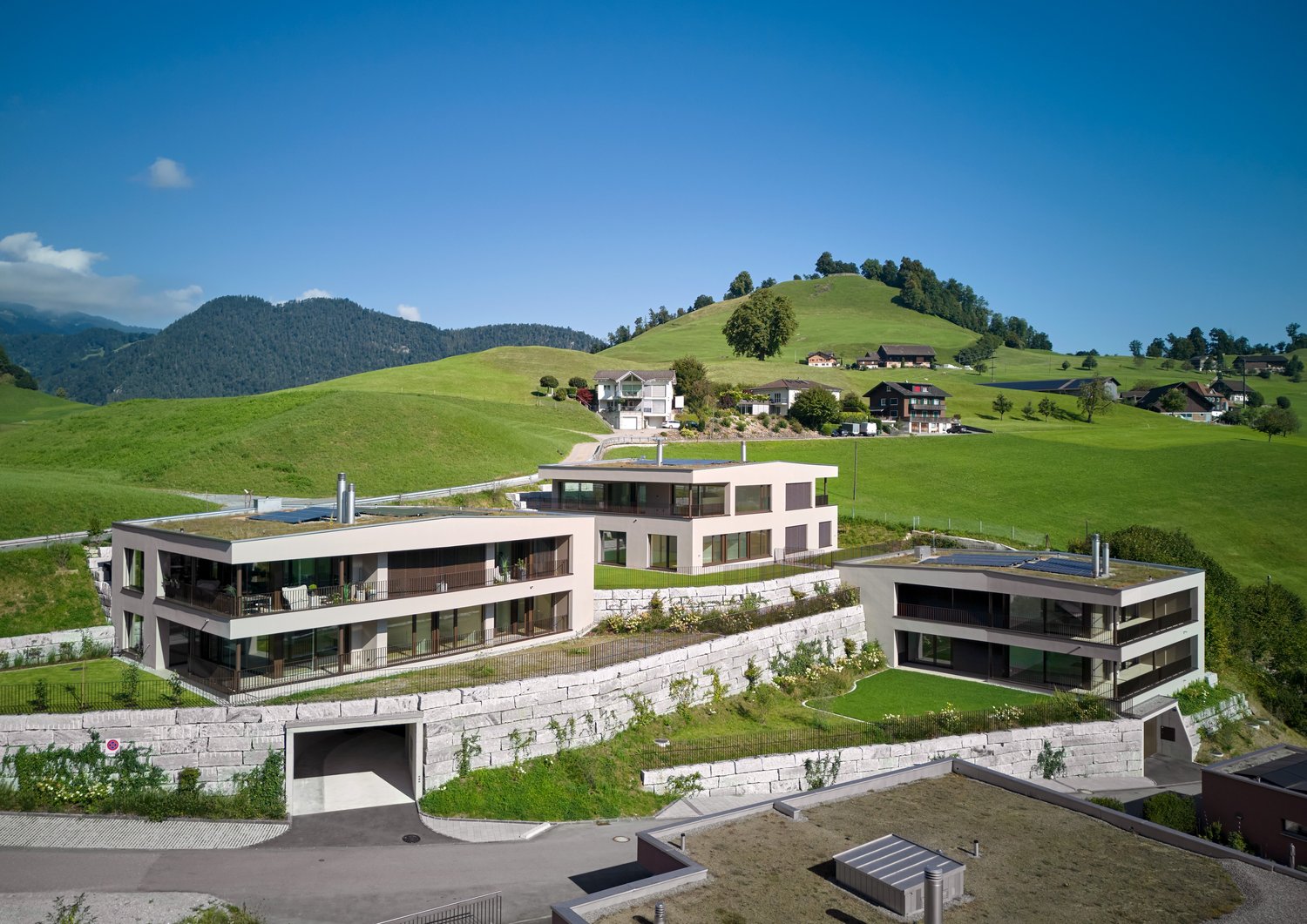 Modern architectural design, multi-story building, white exterior, large windows, solar panels on the roofs, located on a hillside with green surroundings, stone walls, and gardens.
