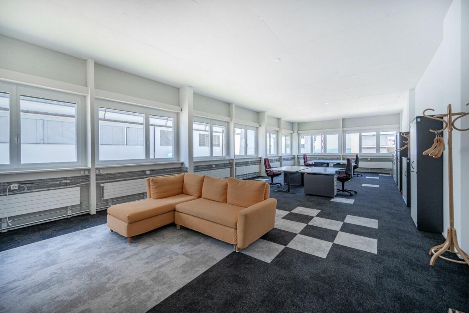 Large office space with a corner sofa, desk, and office chairs, equipped with white radiators and large windows.