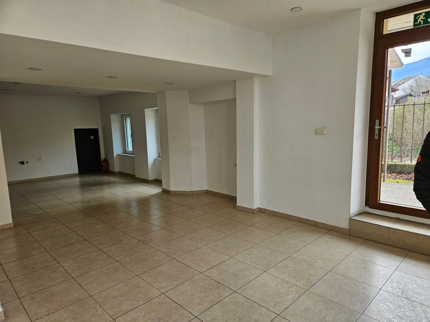 The image shows a large open room with a tiled floor. The walls are painted in a neutral color and there is a window overlooking what appears to be a garden or outdoor area. The room has a simple, minimalist design with no visible furniture or decor.