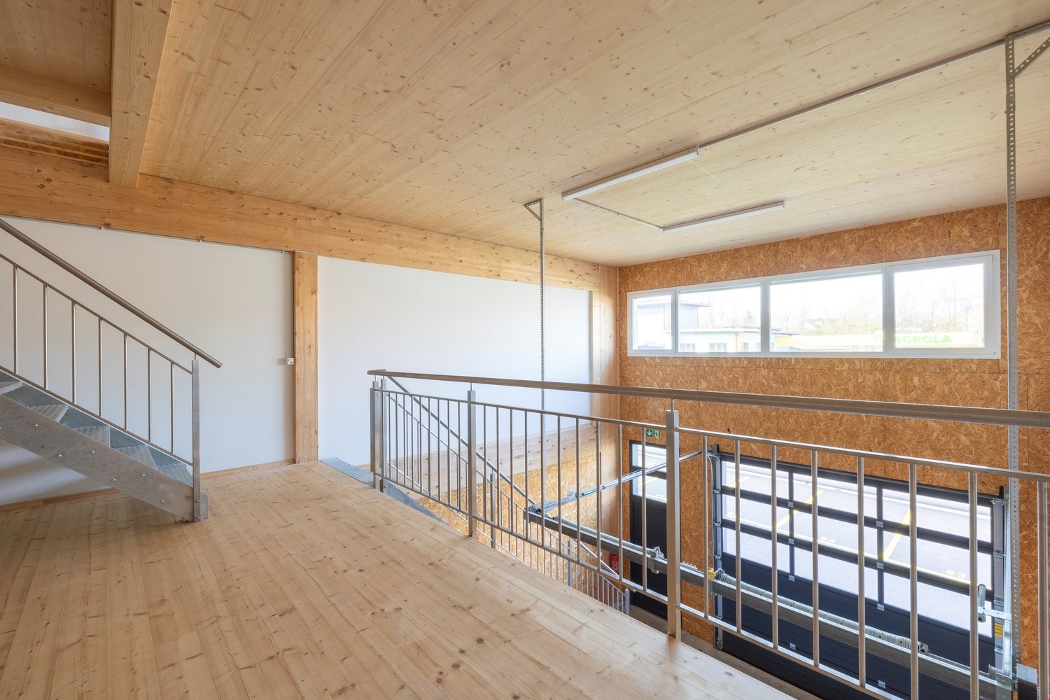 The image shows a large open space with a wooden ceiling and walls. There is a metal railing along the second floor balcony, and the floor is made of wood planks. The room has large windows that provide natural light. The overall design appears to be mode