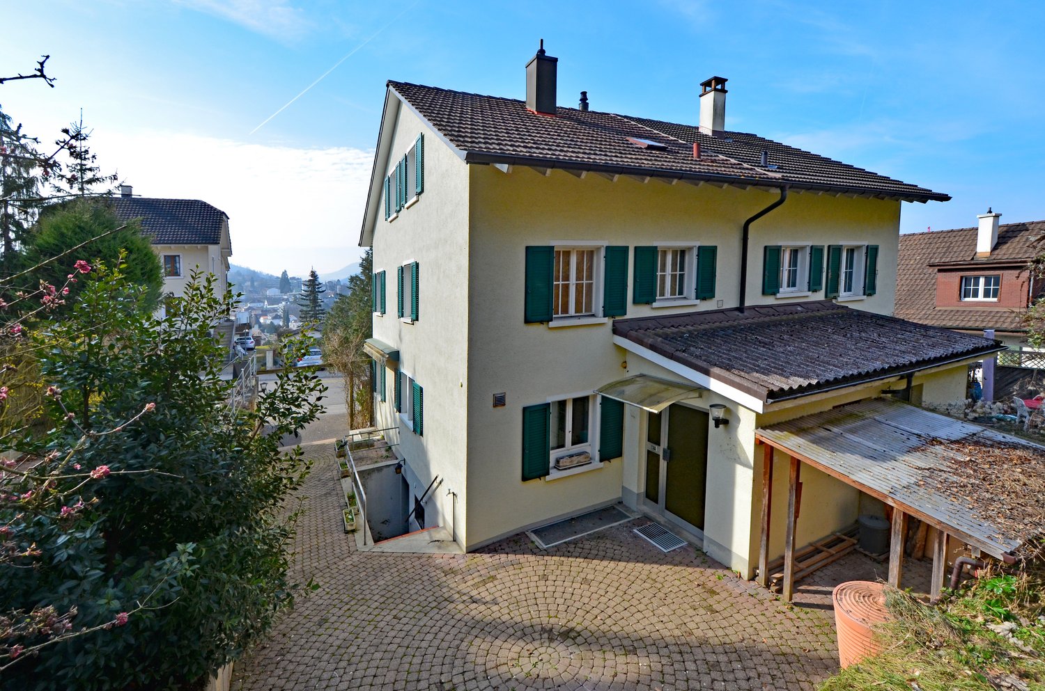 Two-story house with yellow exterior, green shutters, and a tiled roof. The house has a balcony or terrace on the upper level and a paved driveway or walkway leading to the entrance. The surrounding area includes trees and other residential buildings.