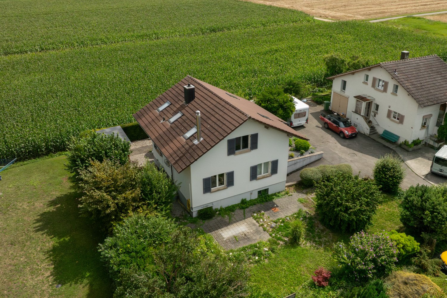 Two detached houses, brown roof on main house, driveway in front, car parked, red car parked by side house, green garden, trees and bushes, field behind